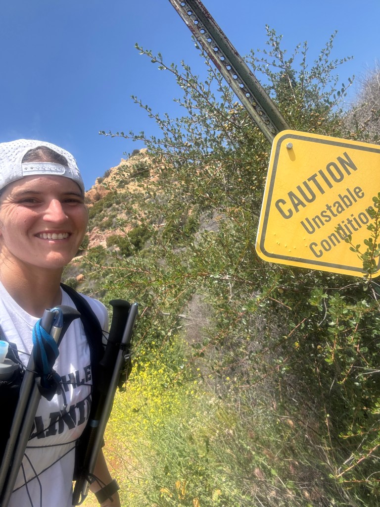 Female hiking on a trail with a sign that says "caution: unstable conditions"